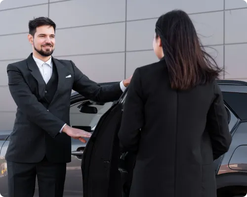 A uniformed chauffeur is holding a car door open for a business executive