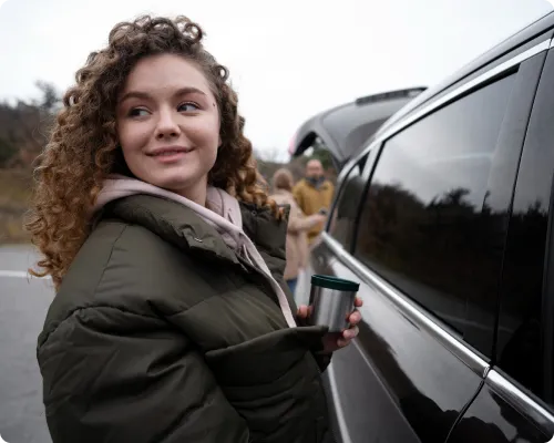 A girl is standing beside a car smiling brightly while her family at the background is getting ready to hop on the car and start their journey