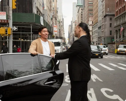 A professional chauffeur is holding a car door open for a client while having a pleasant chat with him