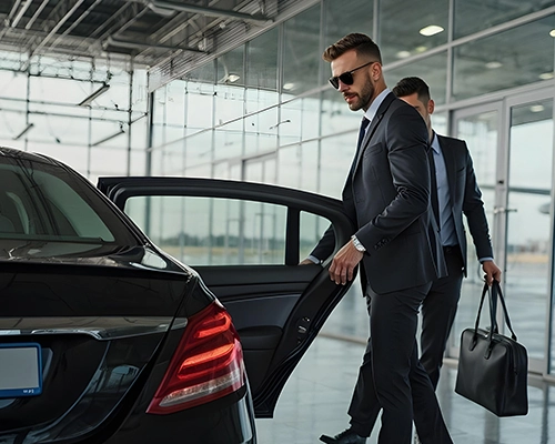 A professional chauffeur in a suit opens the door of a luxury black car at an airport, ready to assist passengers