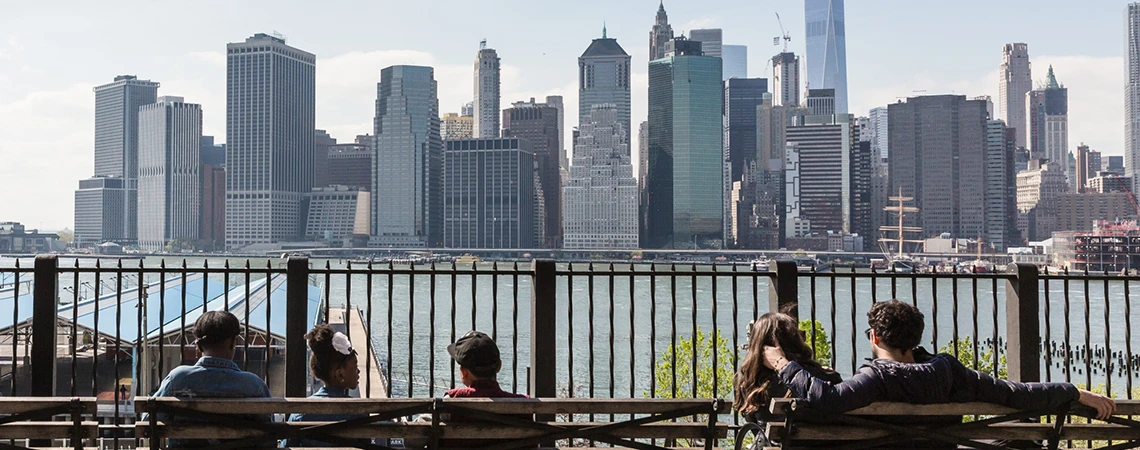The image shows a scenic view of the Manhattan skyline from the Brooklyn Heights Promenade, with people sitting on benches and enjoying the view.