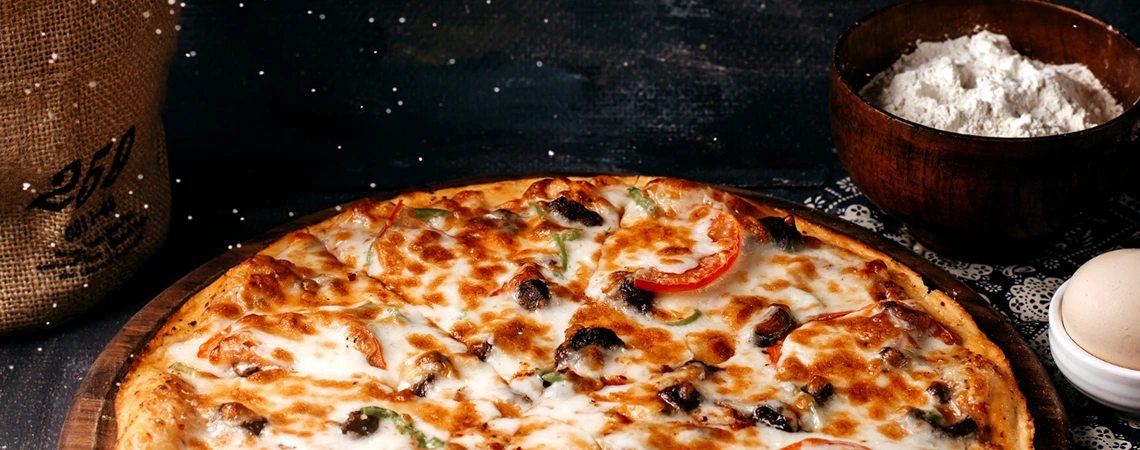 An image of a freshly baked pizza with various toppings on a wooden board, accompanied by a bowl of flour and an egg.
