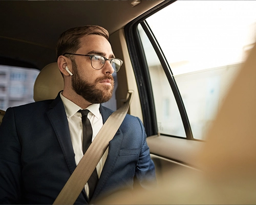 A man in a suit sitting in the back seat of a car, wearing glasses and earbuds, looking out the window.