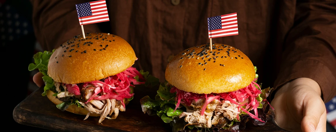Two gourmet burgers topped with American flags are served on a wooden tray, highlighting popular pre-flight meal spots like Ess-a-Bagel in Queens and Massapequa’s All-American Hamburger Drive-In on the way from JFK to Long Island.