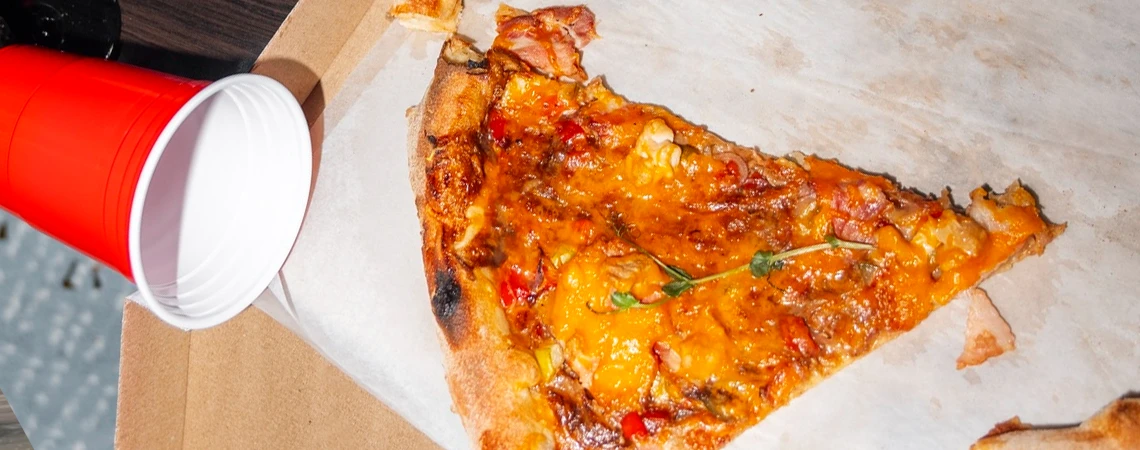 Close-up of a cheesy pizza slice next to takeaway cups, representing quick eats along the New York to Jersey City route with local favorites like La Colombo and Leo’s Grandevous