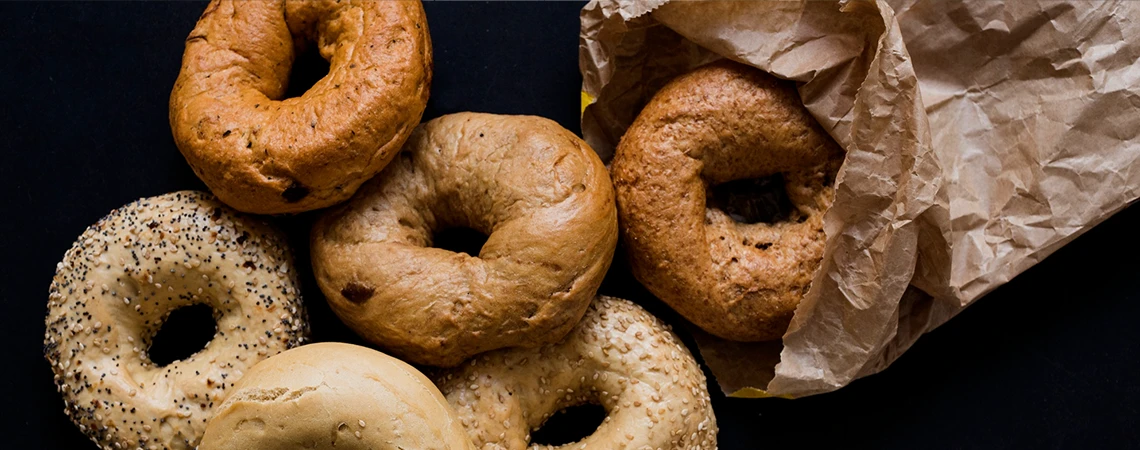 Assorted New York bagels on a dark surface with brown paper bag, promoting quick bite options near JFK before your Philadelphia trip.