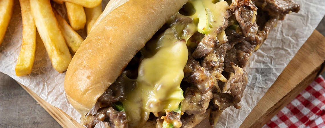 Philadelphia to Newark Airport Car Service Spots for Quick Bite card displaying a close-up of a loaded cheesesteak sandwich with fries, suggesting Pat’s King of Steaks and Federal Donuts for travelers.