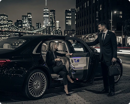 Elegant car service Brooklyn at night: a suited chauffeur assists a stylish woman into a luxury black sedan, with the glowing Brooklyn Bridge and Manhattan skyline in the background.