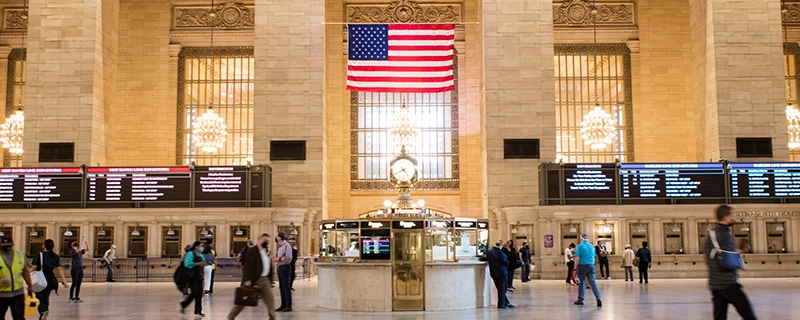 A bustling interior view of Grand Central Terminal with an American flag and architectural details, leading to the American Museum of Natural History. The scene highlights a family-friendly destination with dinosaurs and interactive exhibits, promoting a safe and comfortable journey for kids with child safety seat options and child safety seat availability.