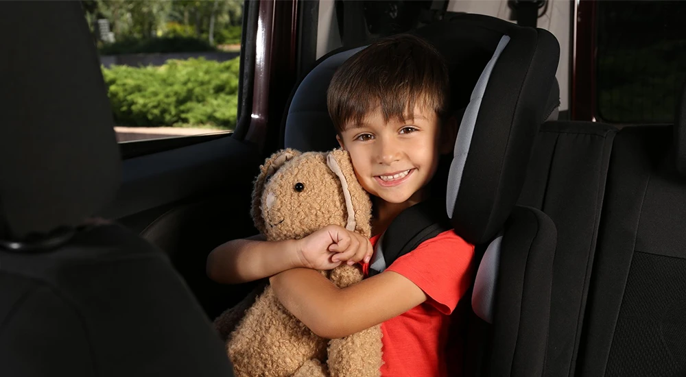 A child seated in a car with a child safety seat, holding a teddy bear, inside a vehicle with a blurred outdoor background. The image accompanies information on New York State Child Safety Regulations, emphasizing safe travel with child safety seats to ensure family safety and compliance on busy NYC roads.