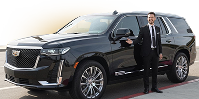 A professional chauffeur in a black suit gesturing toward a luxury black SUV.