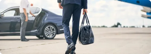 A VIP client is walking towards NYC United limo’s luxury black car while a certified chauffeur is holding the door open for them at JFK airport.
