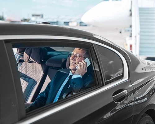 A person in a suit sits in a luxury car at an airport, talking on the phone, highlighting a luxury airport car service offering a personalized, stress-free transfer between JFK and Newark Airport (EWR)