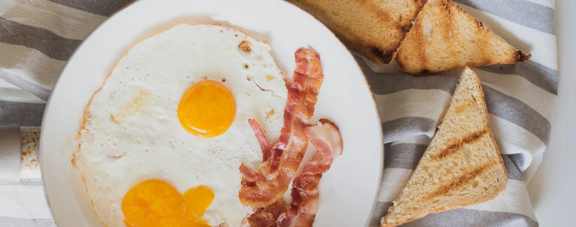 The image shows a plate with a bacon, egg, and cheese sandwich, featuring two sunny-side-up eggs, strips of bacon, and a slice of toast.